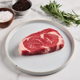 A thick, perfectly marbled Angus Beef Ribeye Steak resting on a dark surface, showcasing its rich red color and intricate fat marbling.
